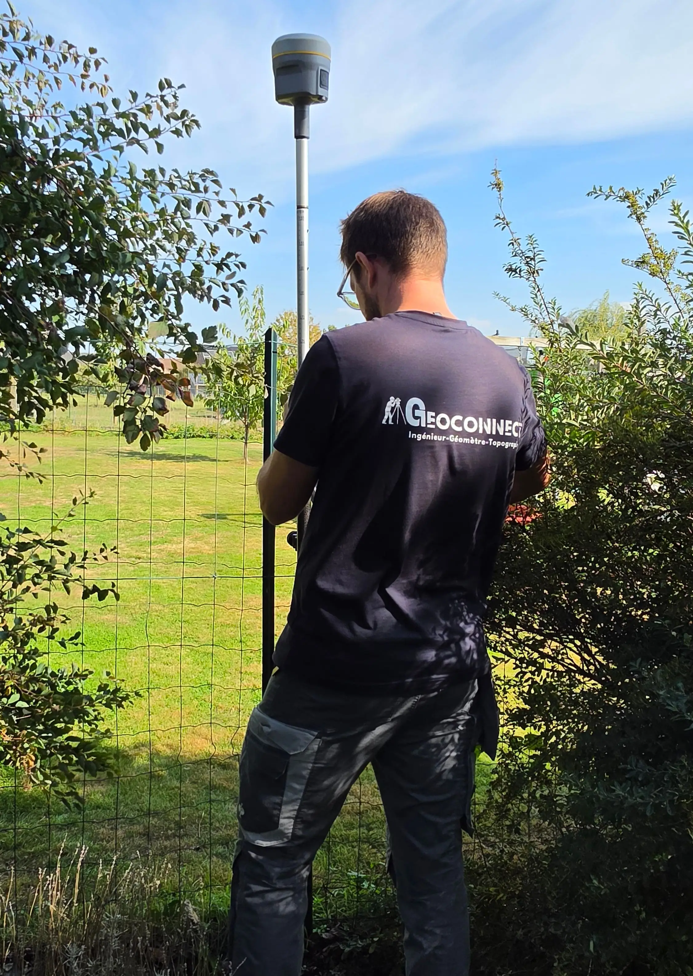 Géomètre de GeoConnect de dos, canne GNSS à la main, le long d’une clôture